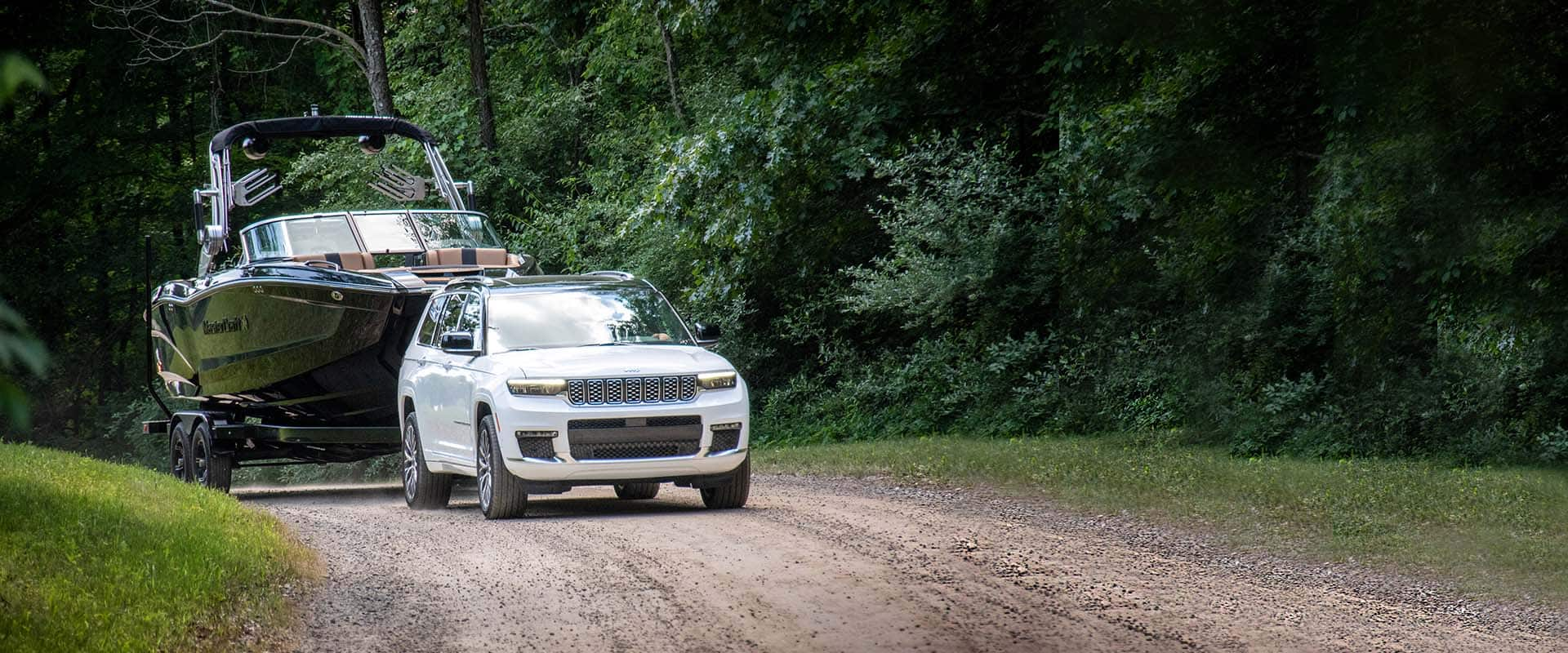 A white 2024 Jeep Grand Cherokee Summit Reserve being driven down a dirt road while towing a motorboat.