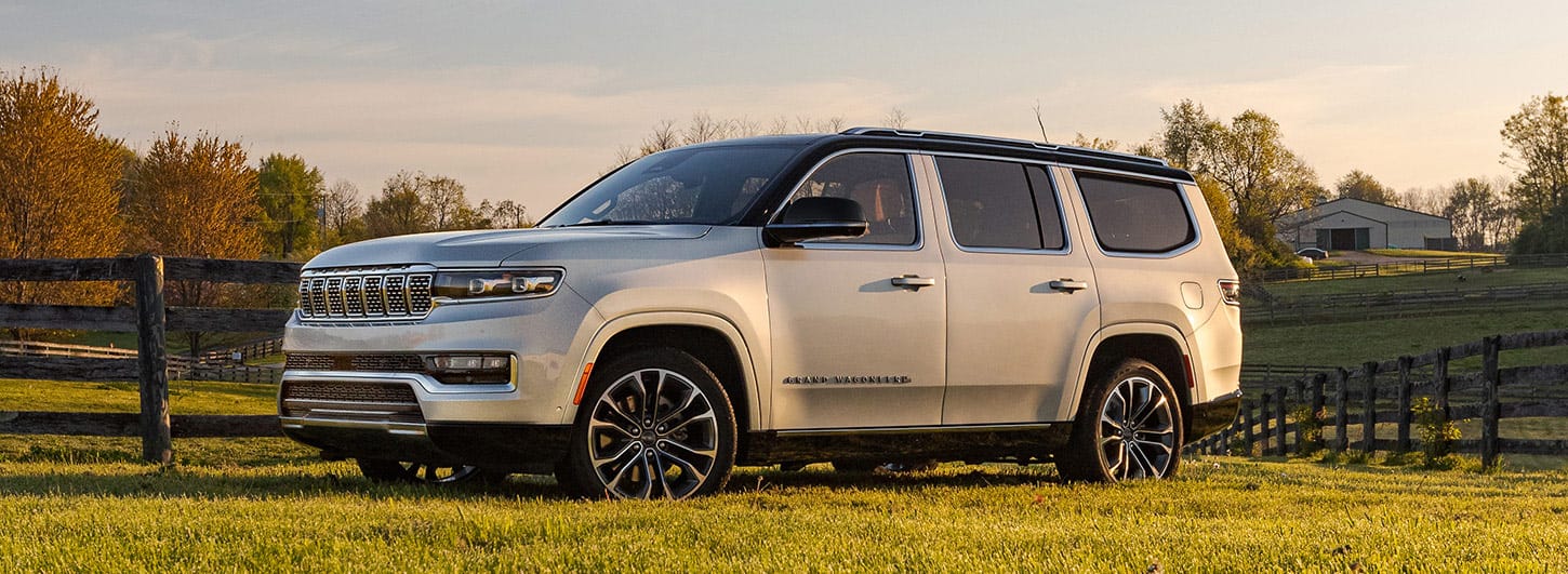 An angled driver-side profile of a beige 2024 Grand Wagoneer Series III parked in a grassy field on a farm at sunset. 