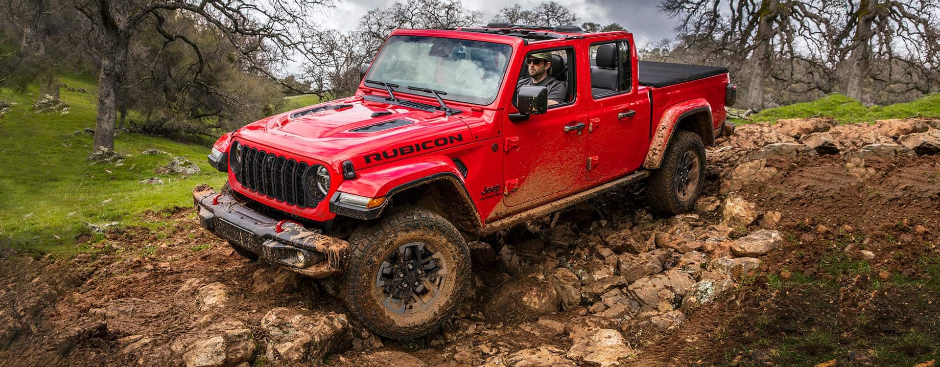 A red 2024 Jeep Gladiator Rubicon with its roof removed, being driven over a rocky, muddy trail off-road.