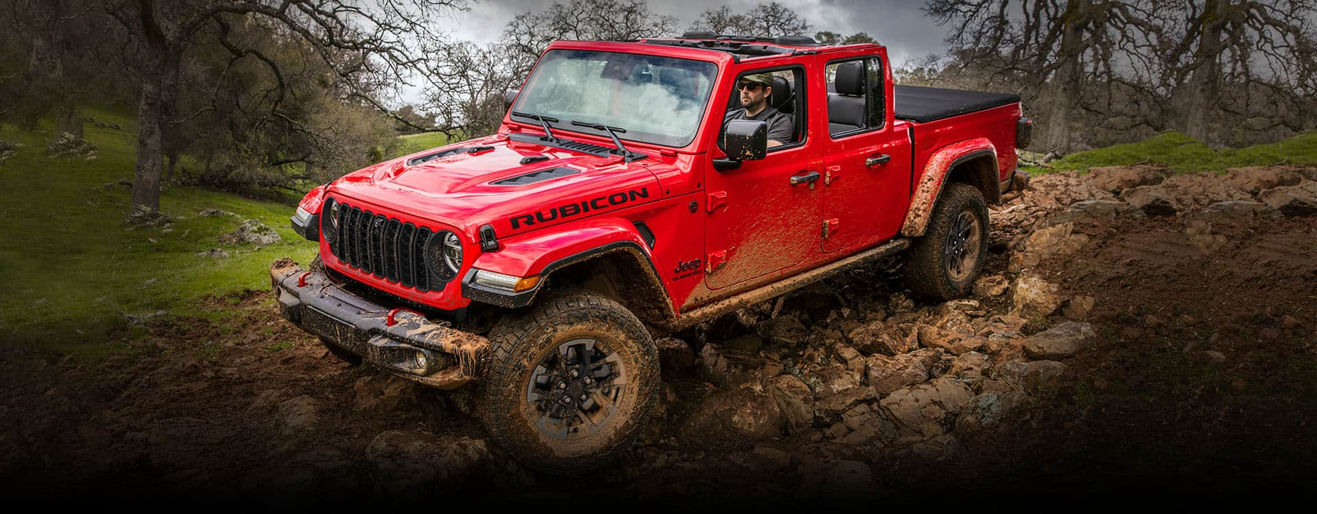 Una Jeep Gladiator Rubicon 2024 roja con el techo desmontado, circulando todoterreno por un sendero rocoso y fangoso.