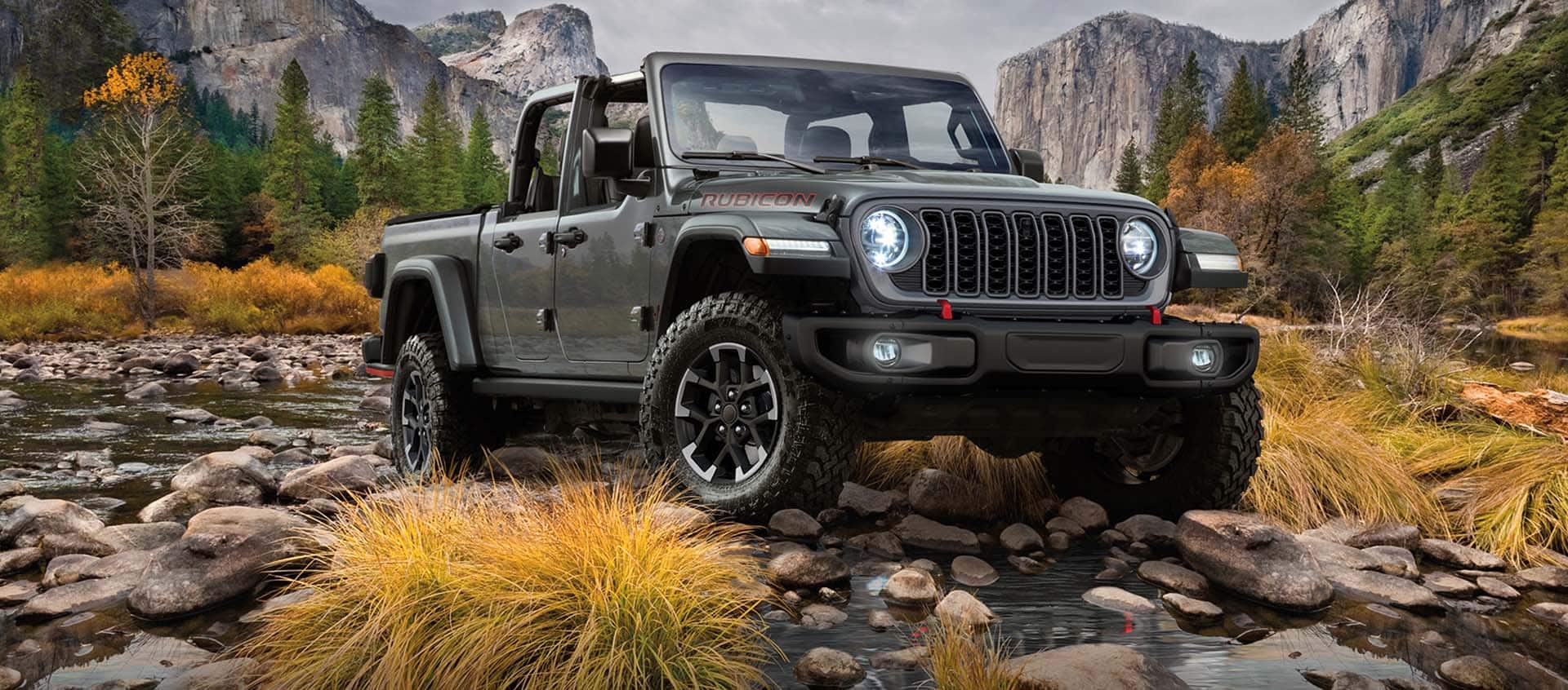 A gray 2024 Jeep Gladiator Rubicon exiting a stream off-road, with mountains in the background.