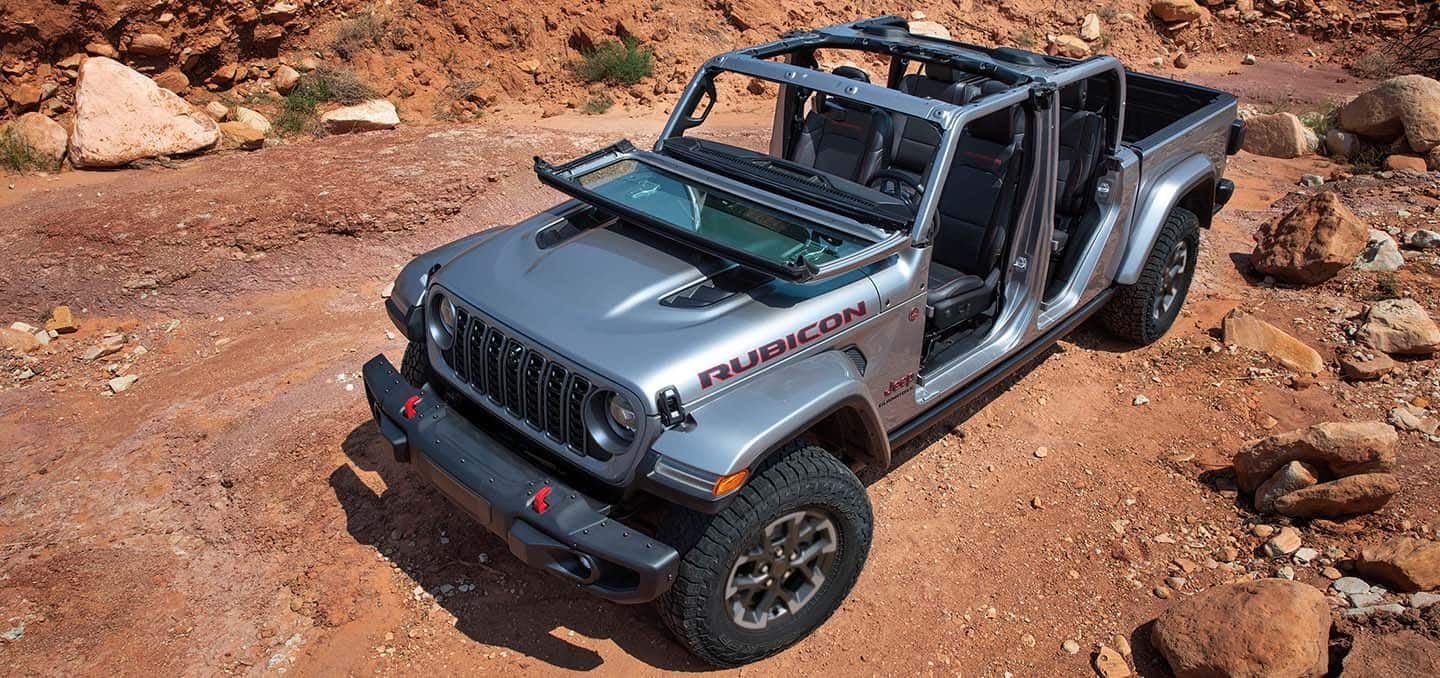 Display A silver 2024 Jeep Gladiator Rubicon with its windshield folded down and doors and roof removed, parked off-road in a clearing.