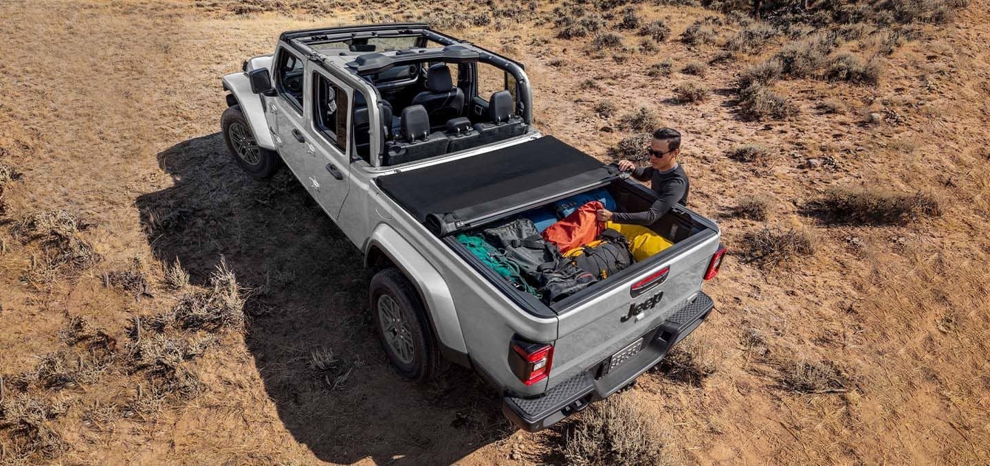 Display A silver 2024 Jeep Gladiator Sport S with its roof removed and tonneau cover partially rolled up to reveal the truck bed packed tightly with camping gear.