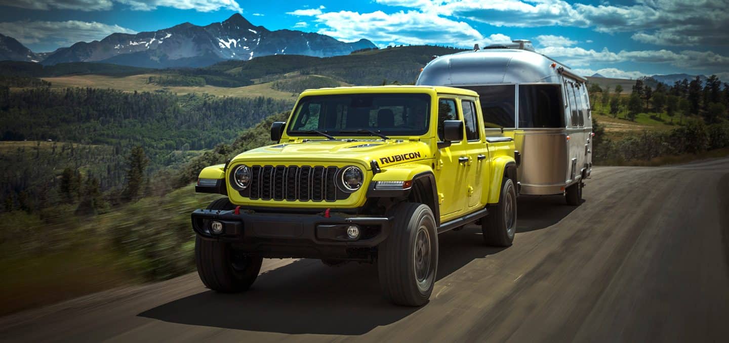 Display A yellow 2024 Jeep Gladiator Rubicon being driven down a highway, towing a travel trailer.