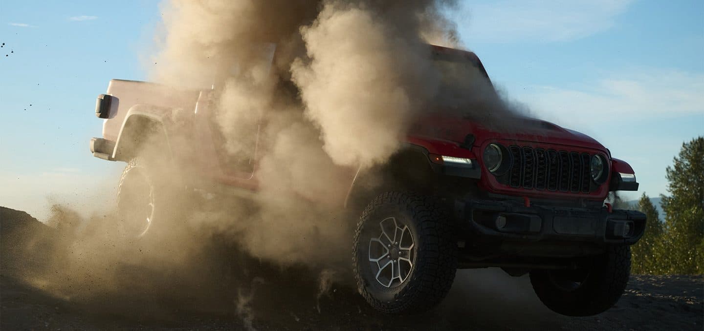 Display A red 2025 Jeep Gladiator pickup truck charging down a hill off-road, with dust nearly engulfing the vehicle.