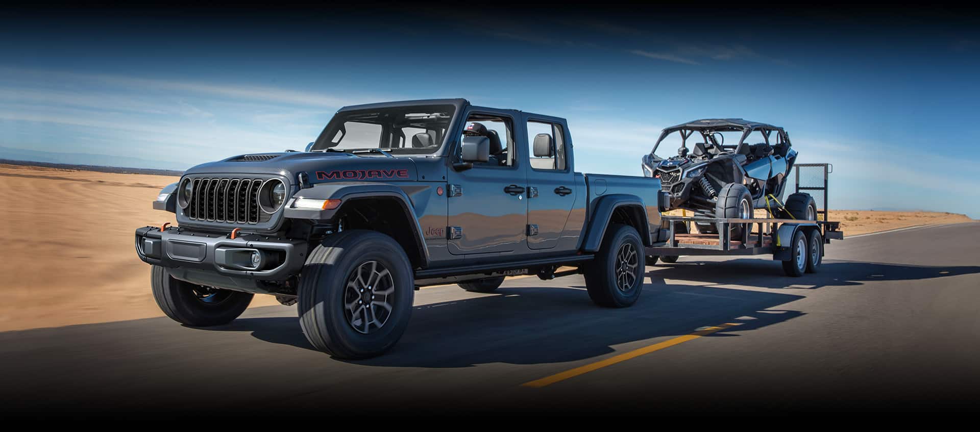 A gray 2024 Jeep Gladiator Mojave with its roof removed, being driven on a highway in the desert, towing an ATV.