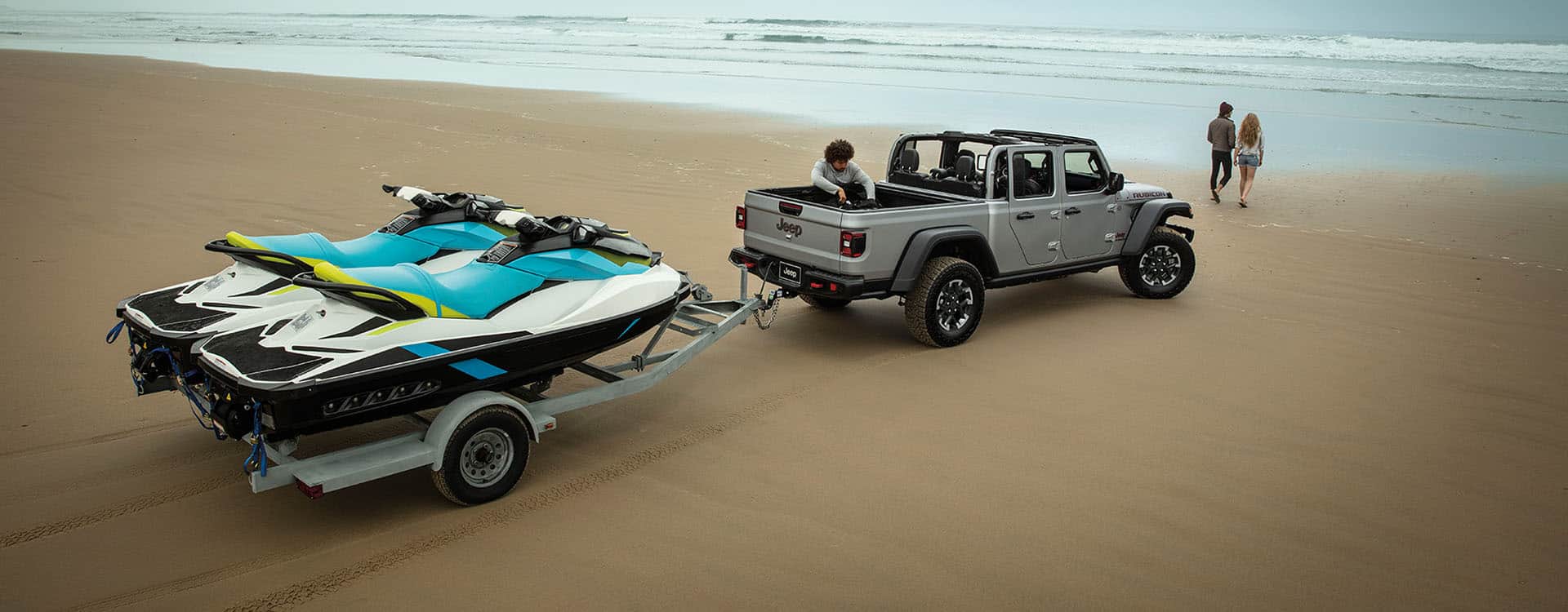 A silver 2024 Jeep Gladiator Rubicon with its roof removed, parked on a sandy beach near the water's edge, towing two personal watercrafts.