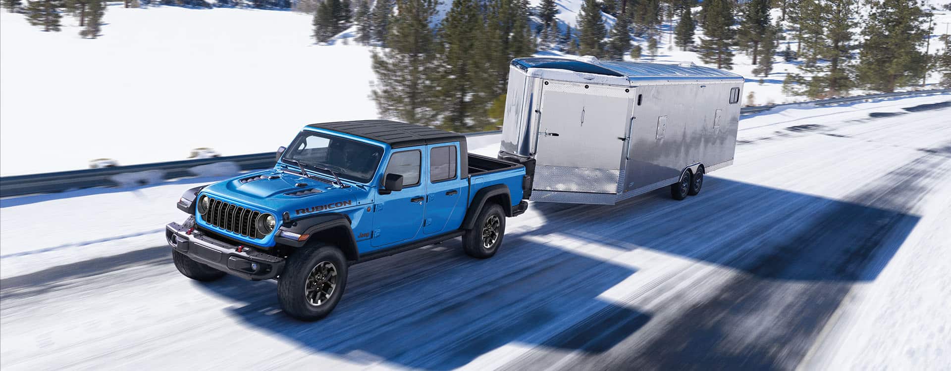 Una Jeep Gladiator Rubicon 2024 azul circulando por una carretera cubierta de nieve mientras acarrea un remolque cerrado.