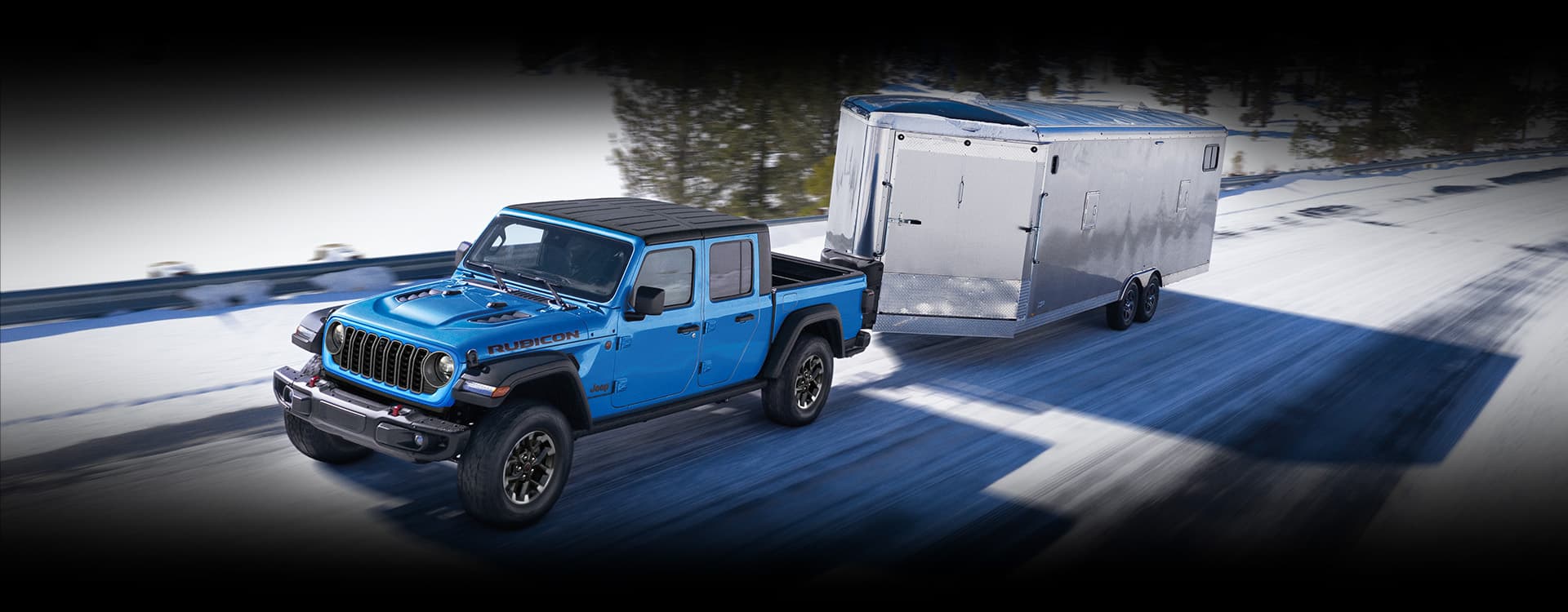 A blue 2024 Jeep Gladiator Rubicon being driven down a snow-covered highway as it tows an enclosed trailer.