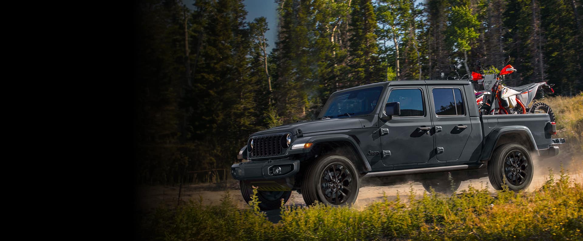 A black 2024 Jeep Gladiator Nighthawk being driven on a trail off-road in the woods with an ATV in its pickup bed.
