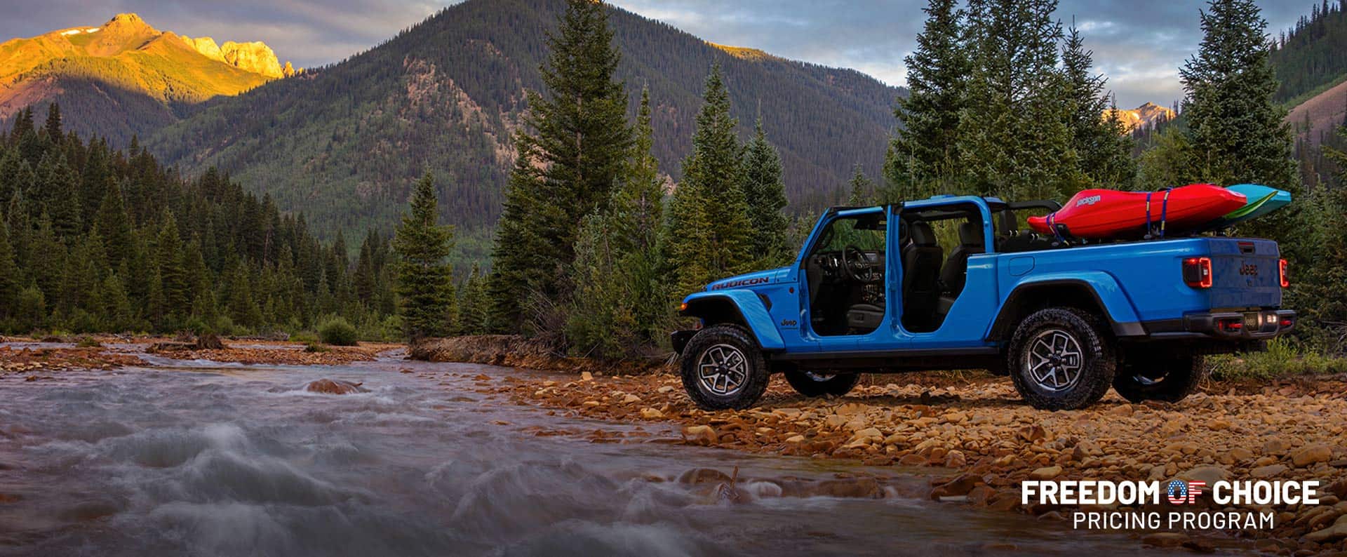 Una Jeep Gladiator Rubicon X 2024 azul estacionada fuera de la carretera, junto a un arroyo rápido entre las montañas. Programa de precios Freedom of Choice.