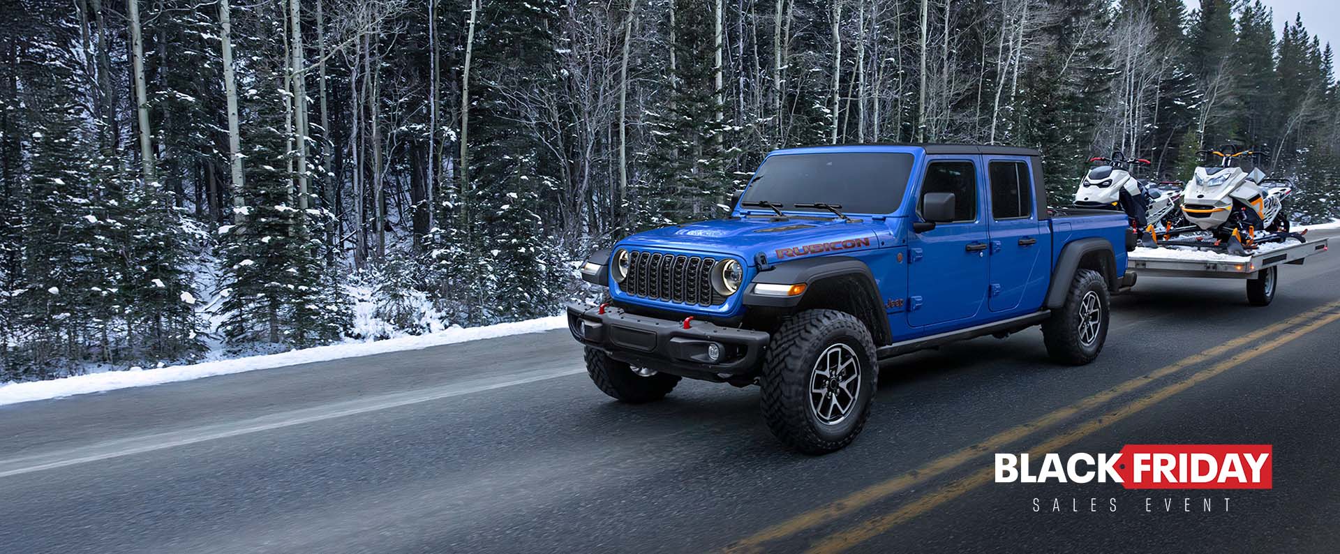 A blue 2024 Jeep Gladiator Rubicon traveling on a highway beside a forest, towing two snowmobiles. The Black Friday Sales Event.