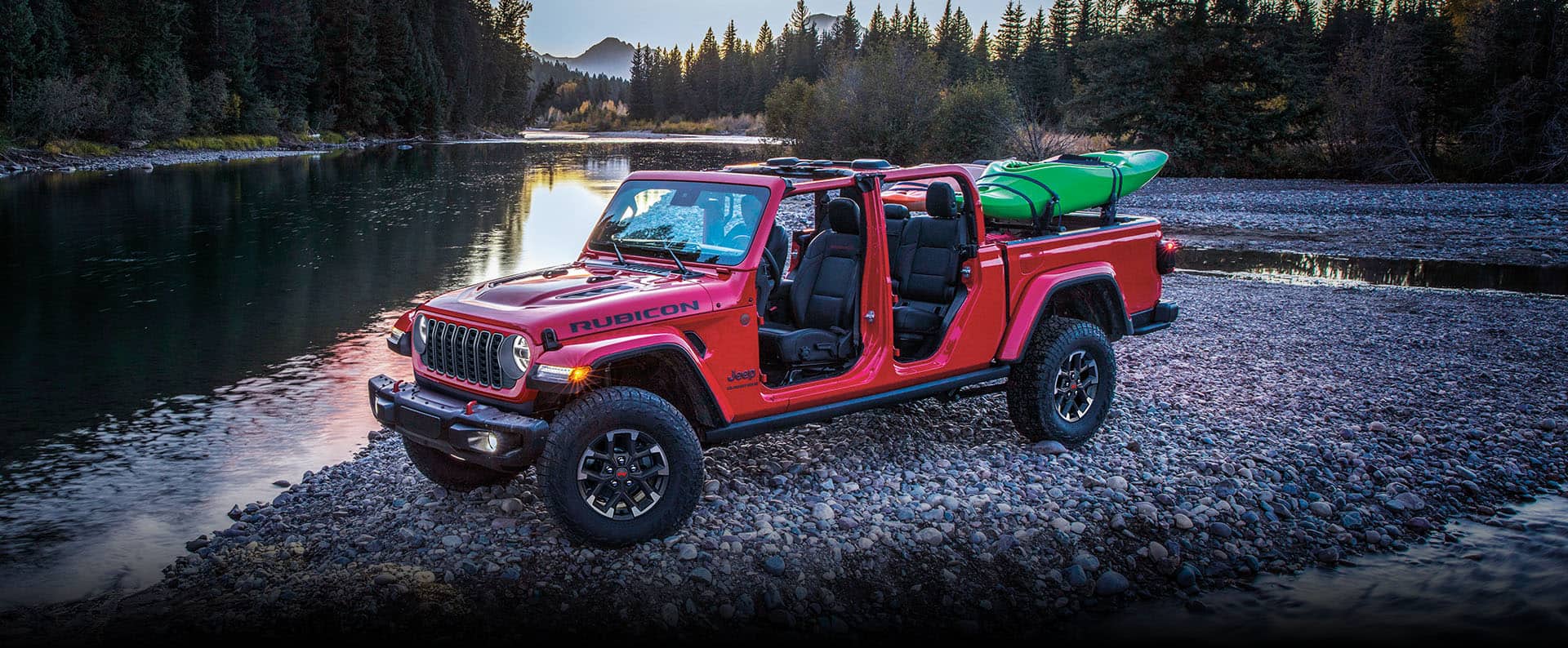 A red 2024 Jeep Gladiator Rubicon X with its roof and doors removed and two kayaks strapped to its pickup box, parked on a mound of rocks surrounded by a stream.