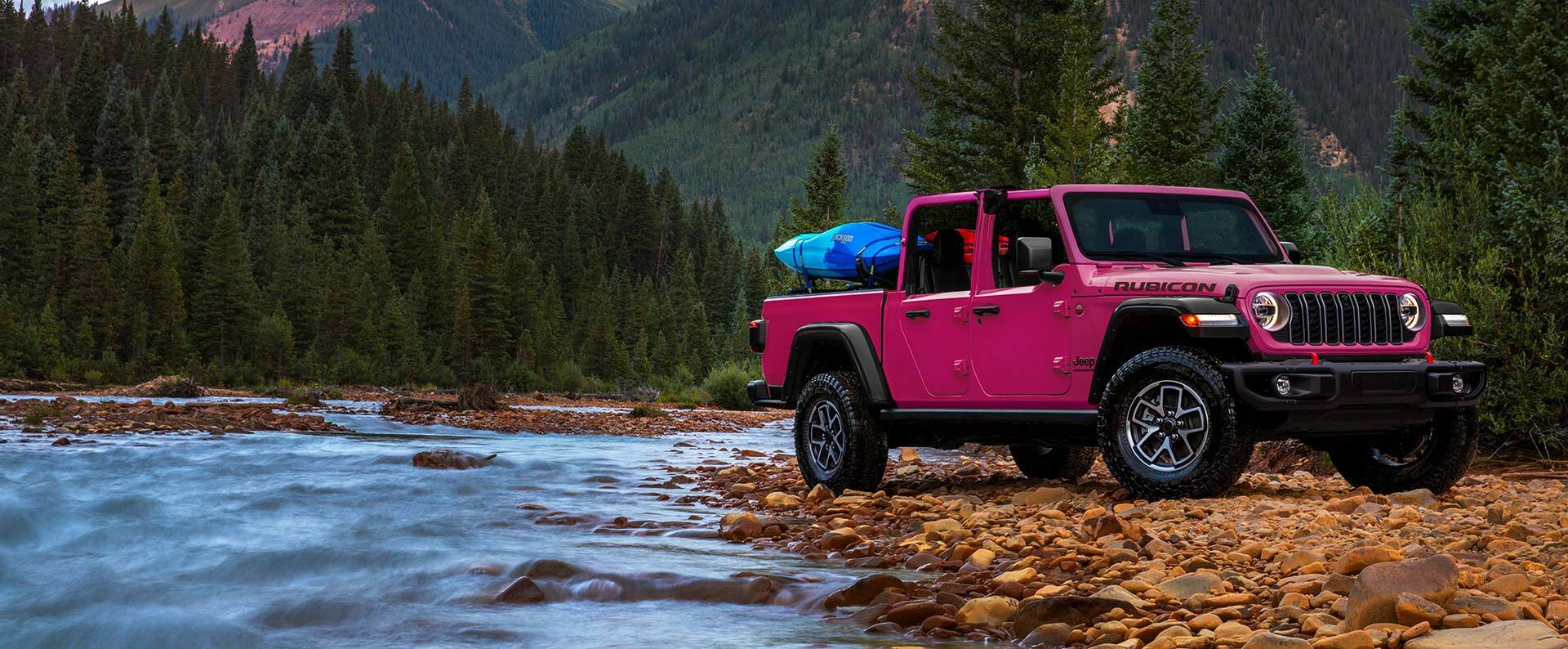 A passenger-side front angle of a pink 2024 Jeep Gladiator Rubicon parked on a bed of rocks beside a stream, with gear in its pickup box and a dense mountain forest in the background.