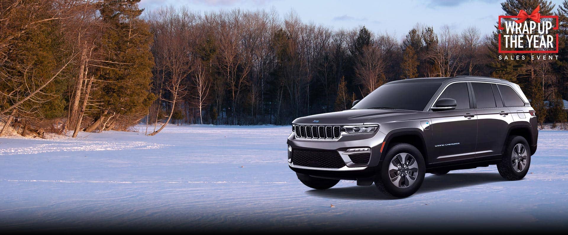 An angled driver-side profile of a gray 2024 Jeep Grand Cherokee 4xe parked on a snow-covered trail, off-road. Wrap Up The Year Sales Event.