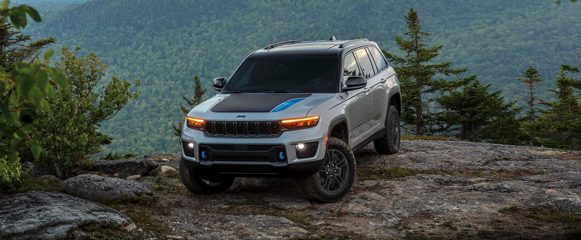A silver 2024 Jeep Grand Cherokee Trailhawk with black hood insert, parked on the edge of a cliff in the mountains.
