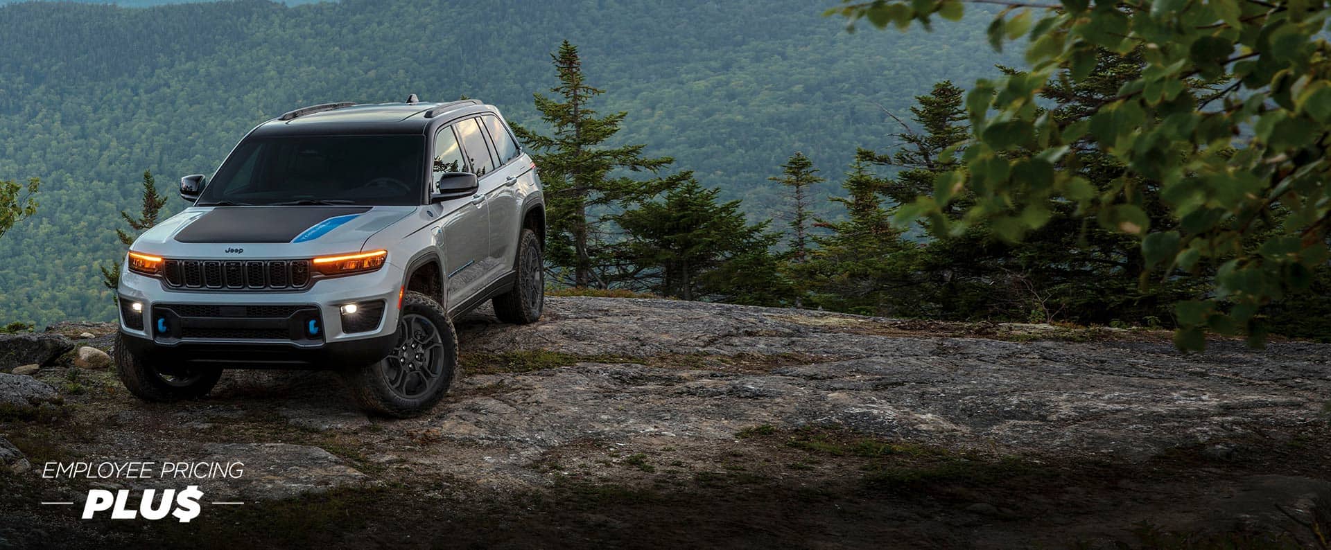 A silver 2024 Jeep Grand Cherokee Trailhawk with black hood insert, parked on the edge of a cliff in the mountains. Employee Pricing Plus. 