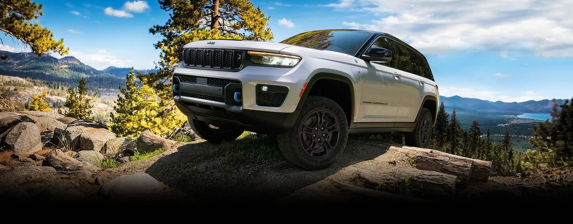 A white 2024 Jeep Grand Cherokee Trailhawk 4xe with black hood insert, being driven over an outcropping, with mountains and a lake in the background.