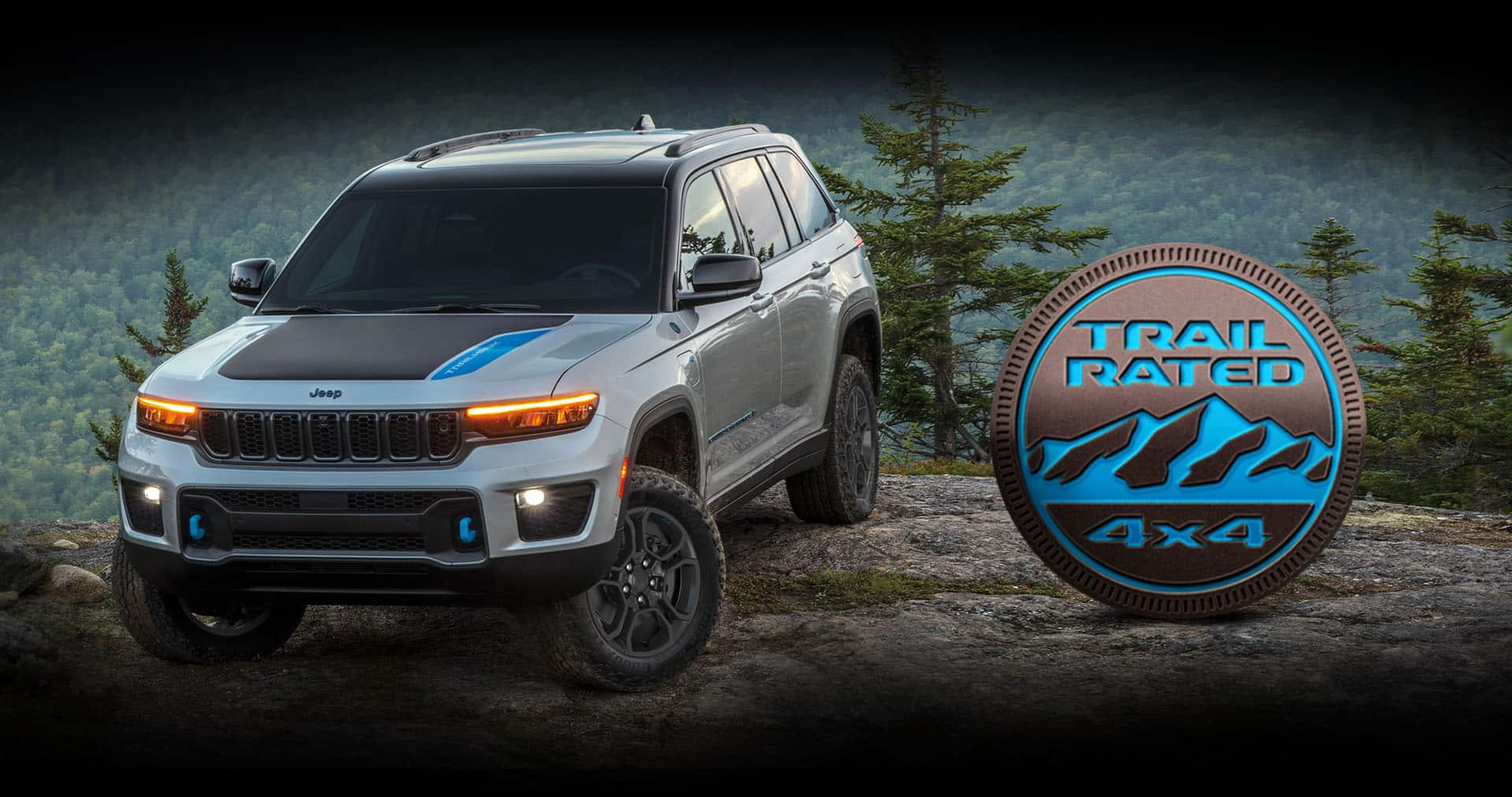 A silver 2024 Jeep Grand Cherokee Trailhawk 4xe parked on a clearing off-road, with a mountain forest in the background. The Trail Rated 4x4 badge.