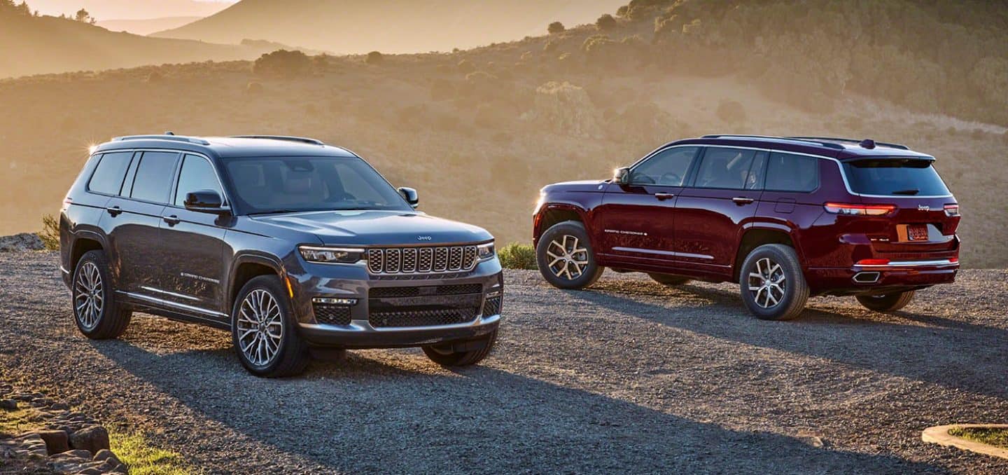 Display A gray 2024 Jeep Grand Cherokee Summit Reserve and a red Jeep Grand Cherokee Limited parked opposite of each other on a gravel mountain road at sunset.