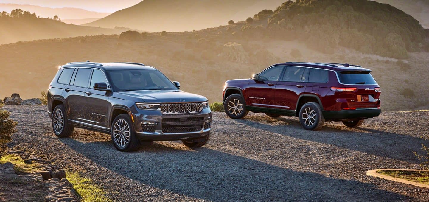 Display A gray 2024 Jeep Grand Cherokee Summit Reserve and a red Jeep Grand Cherokee Limited parked opposite of each other on a gravel mountain road at sunset.