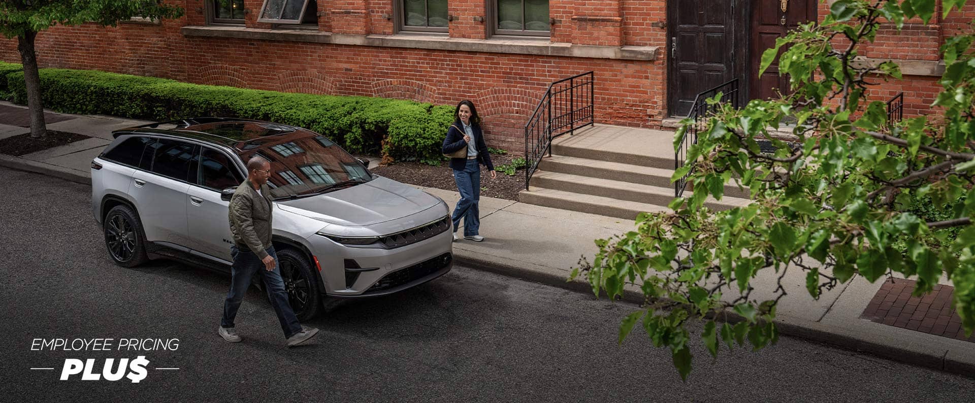 A silver 2024 Jeep Wagoneer S Launch Edition parked beside a brick building with a man walking by the passenger side and a woman walking by the driver side. Employee Pricing Plus.