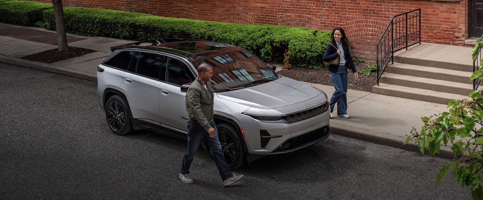 A silver 2024 Jeep Wagoneer S Launch Edition parked beside a brick building with a man walking by the passenger side and a woman walking by the driver side.