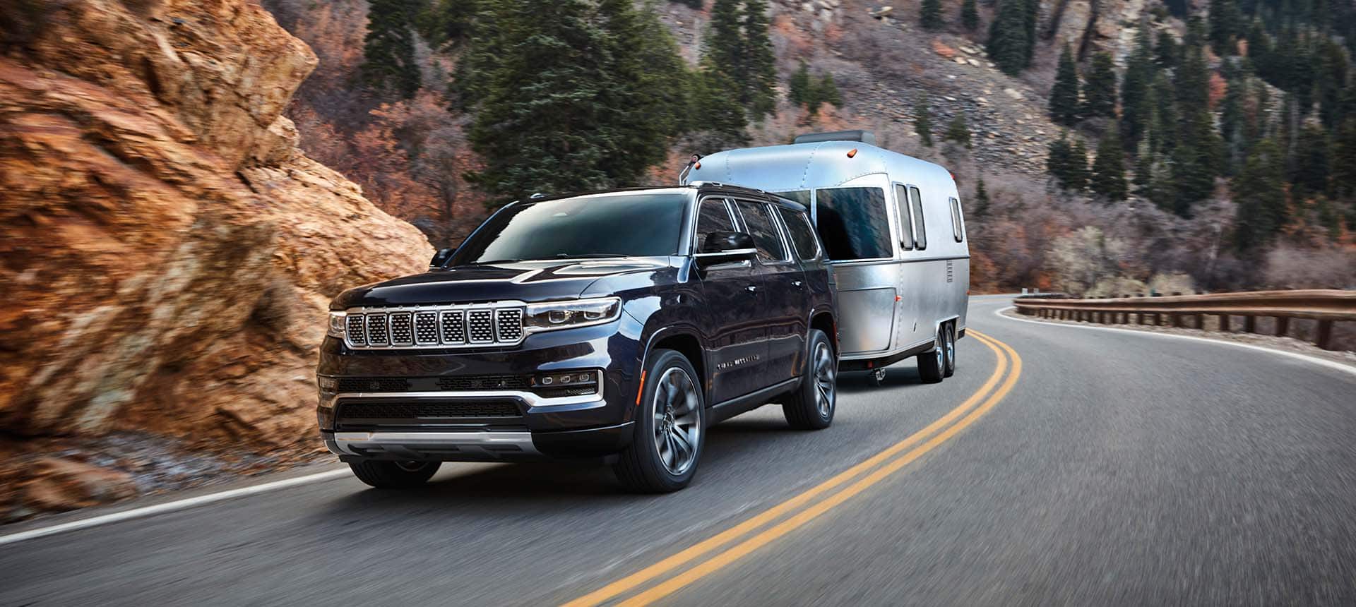 A black 2024 Grand Wagoneer Series III towing a travel trailer as it is driven down a winding highway in the mountains.