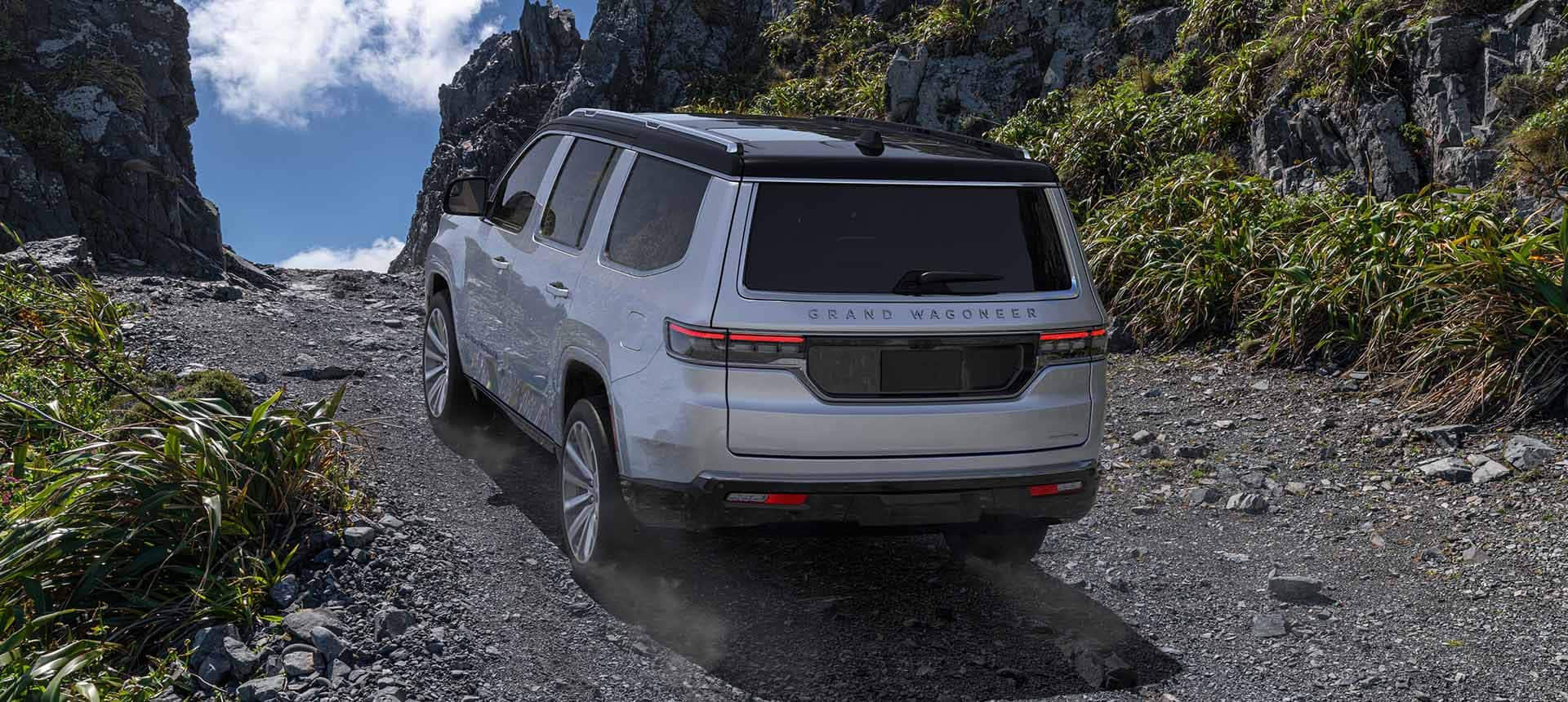 A rear angle of a silver 2024 Grand Wagoneer Series III ascending a hill off-road, surrounded by grass-covered rock faces.