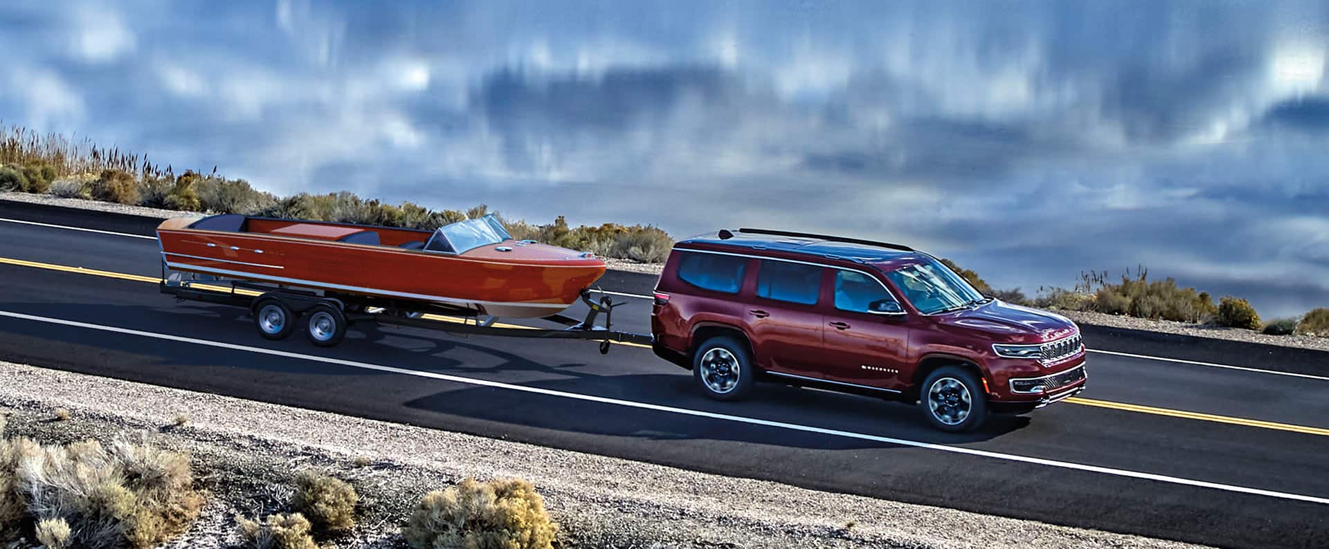 The 2024 Wagoneer Series III towing a motorboat on a lakeside road, with storm clouds overhead.
