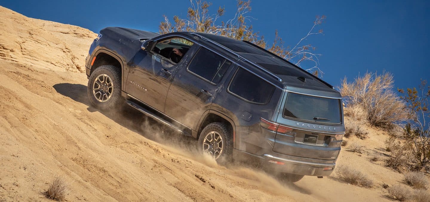 Display A gray 2024 Wagoneer Series III ascending a steep, sandy hill off-road.