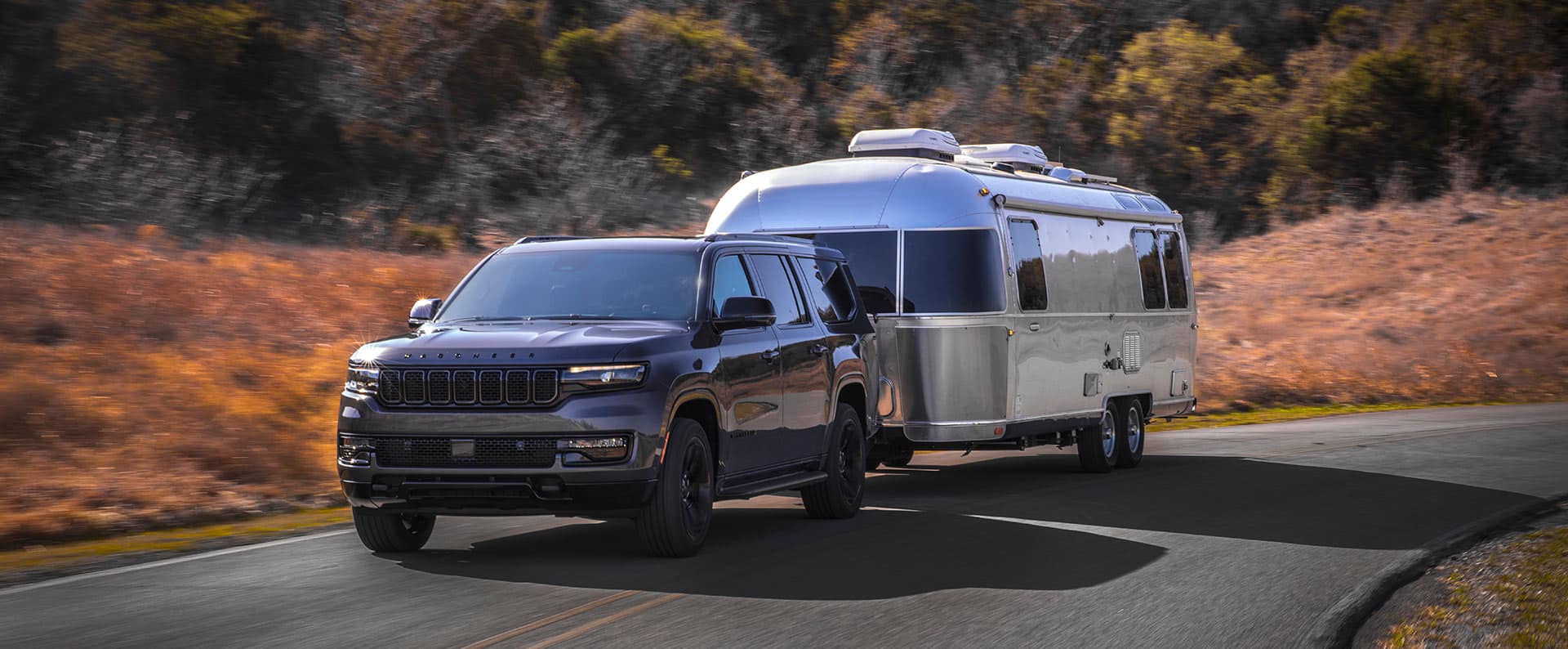 A black 2024 Wagoneer Carbide towing a travel trailer as it is driven down a rural highway.