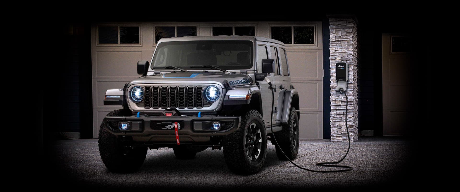 A 2024 Jeep Wrangler Rubicon 4xe parked in a garage with a charging station on the wall and the charging cord plugged into the vehicle's charging port.