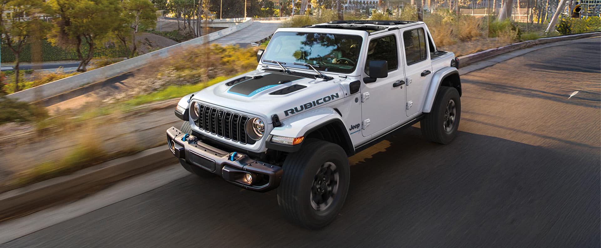 Un Jeep Wrangler Rubicon X 4xe 2024 blanco con una inserción negra en el capó, circulando por un calle con edificios altos en el fondo.