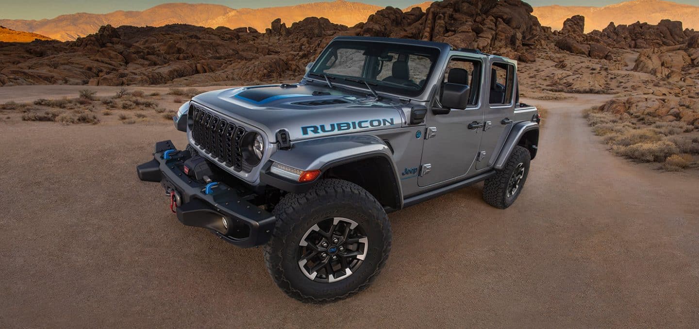 Display An angled driver-side profile of a gray 2024 Jeep Wrangler Rubicon 4xe parked on a clearing in the desert.