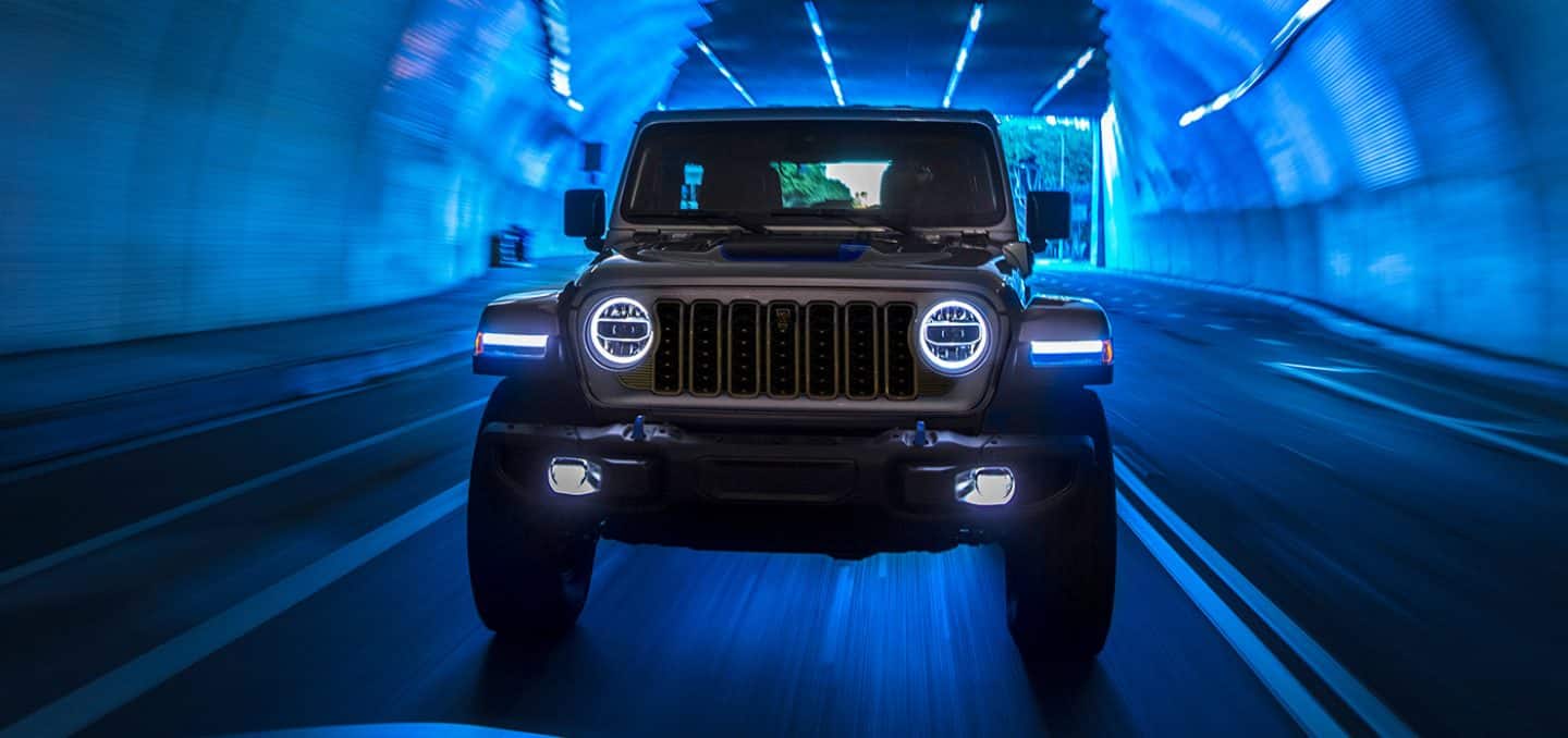 Display A front angle of a gray 2024 Jeep Wrangler Rubicon 4xe with its headlamps on, being driven through a lighted tunnel, with the entire image tinted blue.
