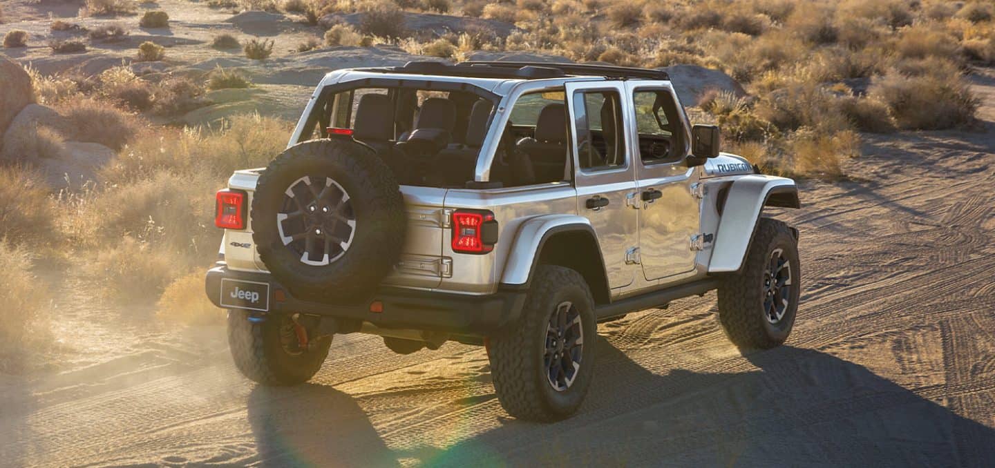 Display A rear angle of a white 2024 Jeep Wrangler Rubicon 4xe being driven on a sandy trail, off-road in the desert.
