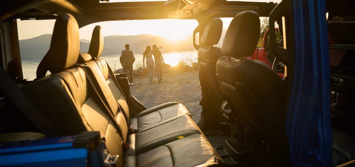 Display The rear seat in the 2025 Jeep Wrangler with its doors removed and three people walking away from the vehicle, toward a lake, with mountains in the background at sunrise.