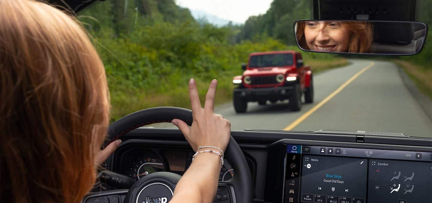Display The interior of a 2025 Jeep Wrangler with a female driver giving the Jeep Wave (raising the first two fingers on her right hand) to the driver of a red 2025 Jeep Wrangler traveling on the same highway, in the opposite direction, displayed through the windshield.