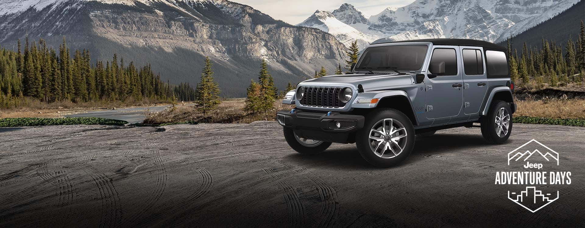 A gray 2024 Jeep Wrangler Sport S 4xe parked on a clearing beside a river, with mountains in the background. Jeep Adventure Days logo.