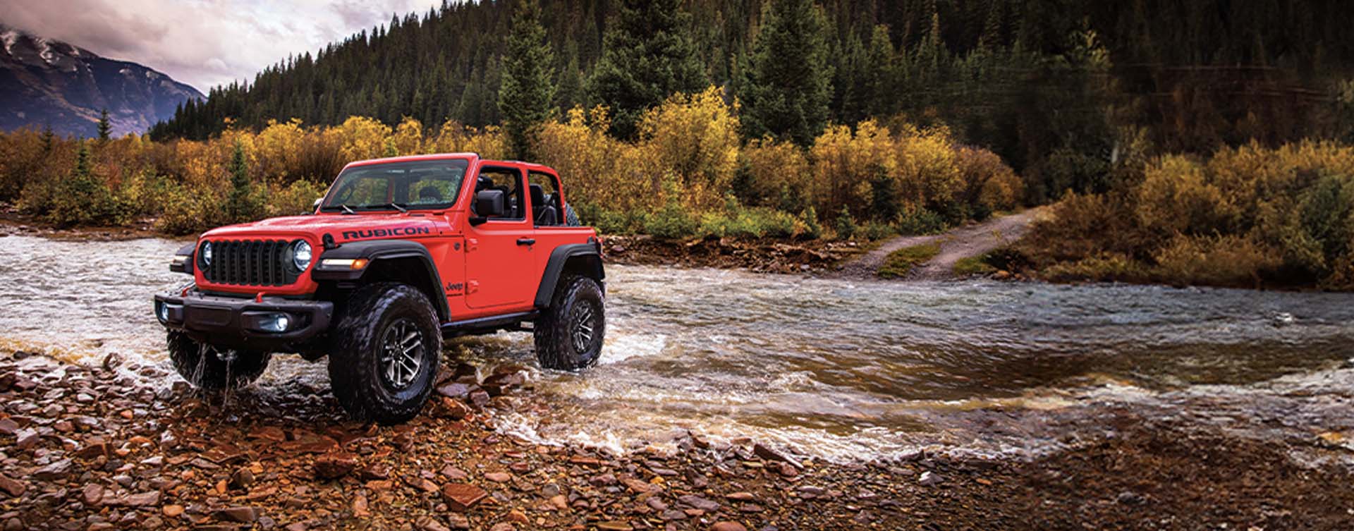 A red 2024 Jeep Wrangler Rubicon two-door with the XTreme 35 Tire Package and its roof removed, fording through a stream off-road, with a thick forest mountain in the background.