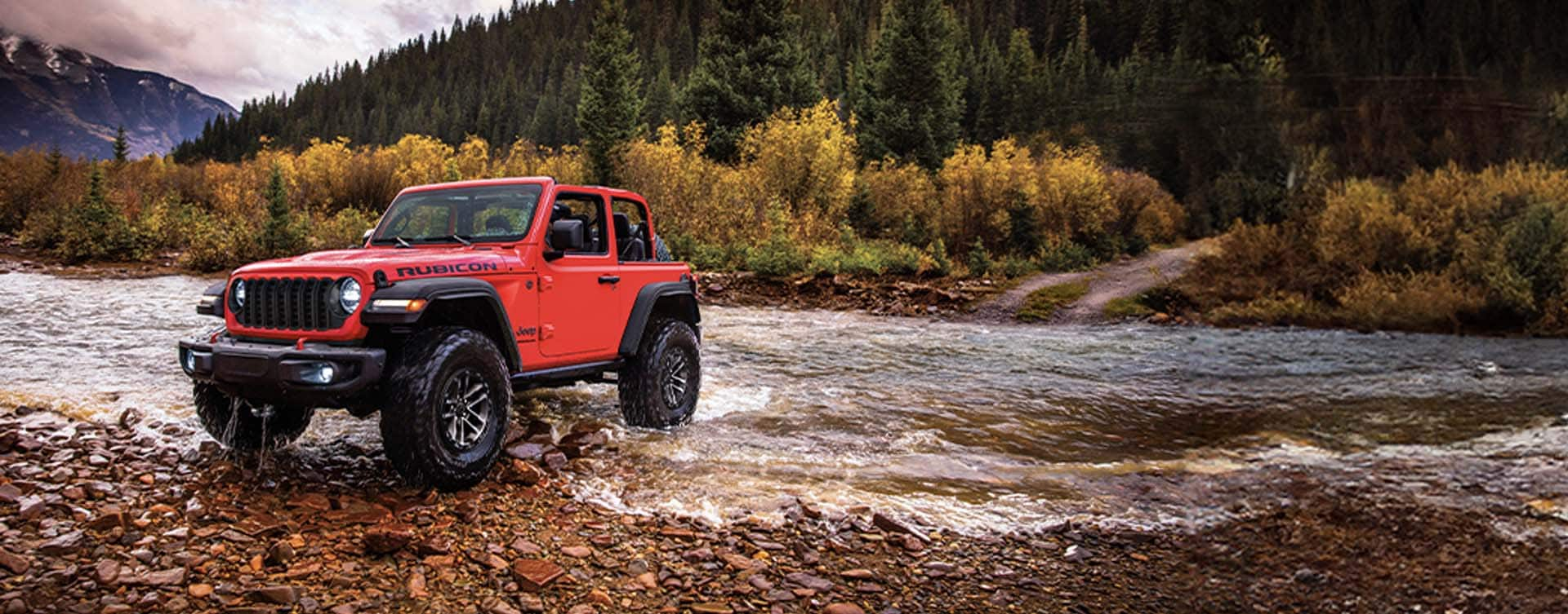 A red 2024 Jeep Wrangler Rubicon two-door with the XTreme 35 Tire Package and its roof removed, fording through a stream off-road, with a thick forest mountain in the background.