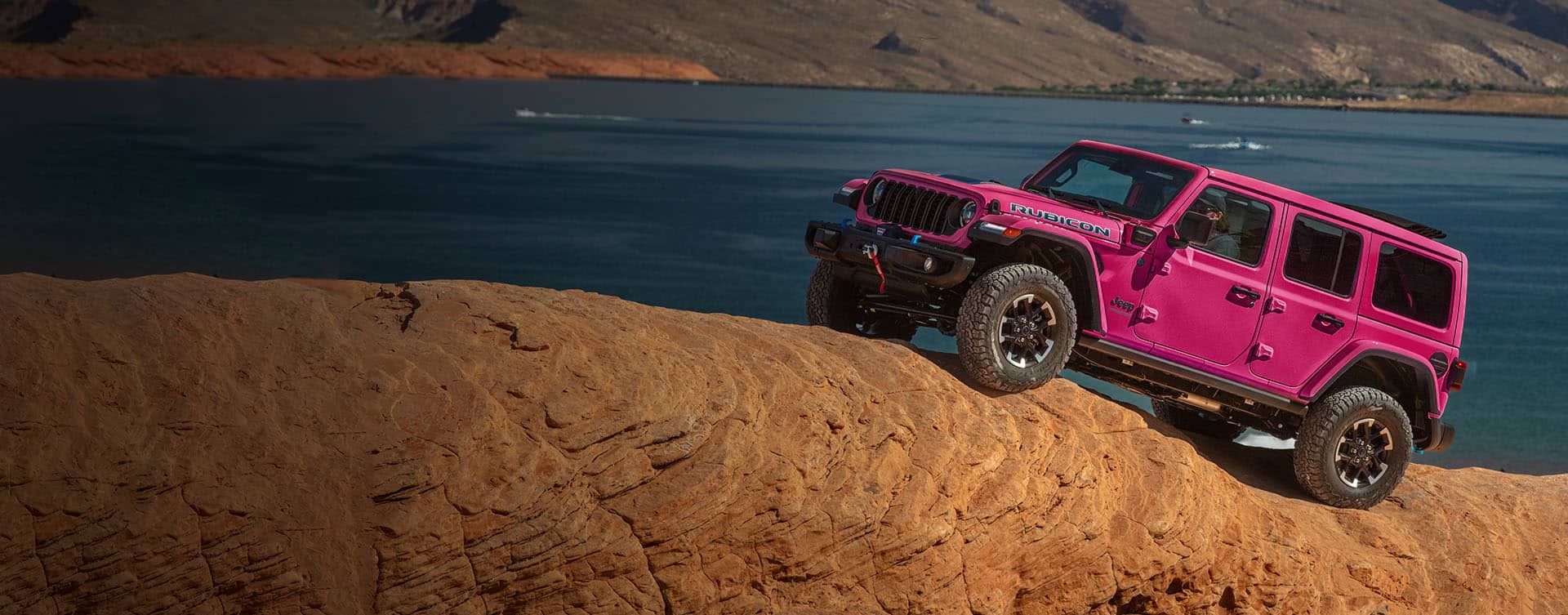 A hot pink 2024 Jeep Wrangler Rubicon four-door ascending a steep, rock hill off-road, with a body of water in the background.