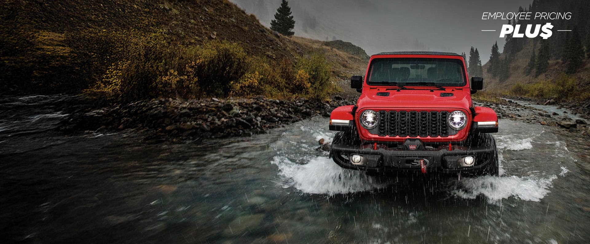 Ángulo frontal de un Jeep Wrangler Rubicon 2024 rojo que circula a través de un arroyo, siguiendo su camino. Evento de precios de empleado.