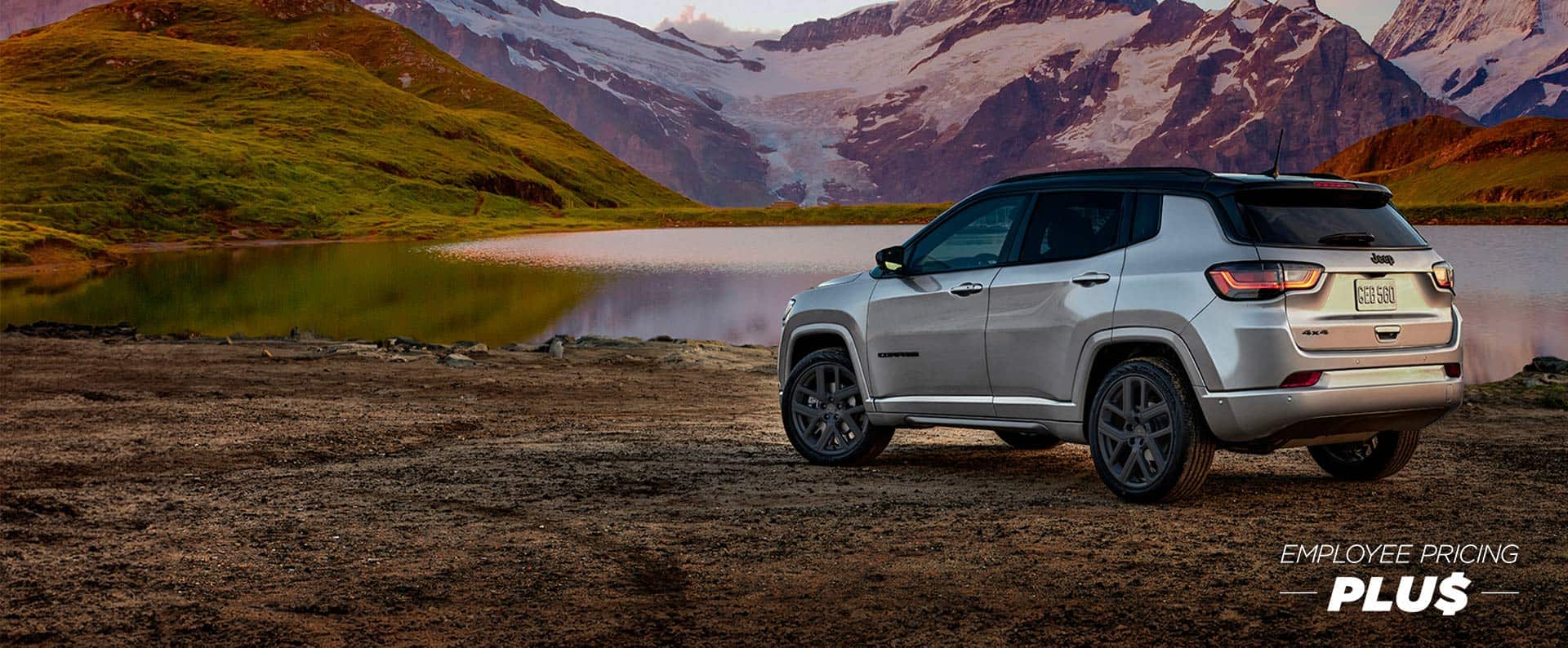 A silver 2025 Jeep Compass Limited with the High Altitude Package, parked beside a mountain lake at sunset. Employee Pricing Plus event.