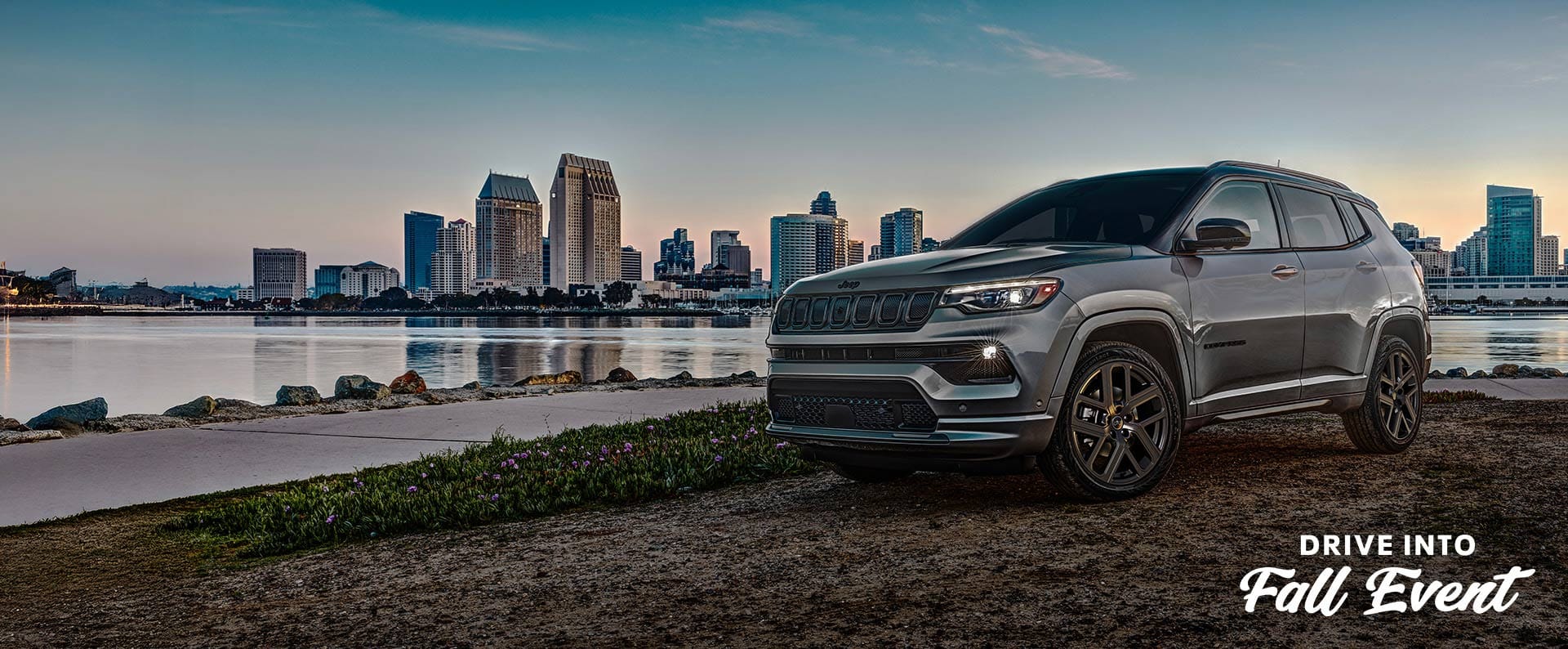 A driver-side front angle of a gray 2025 Jeep Compass Limited High Altitude parked at the riverfront, with tall building across the water. Drive Into Fall Event.