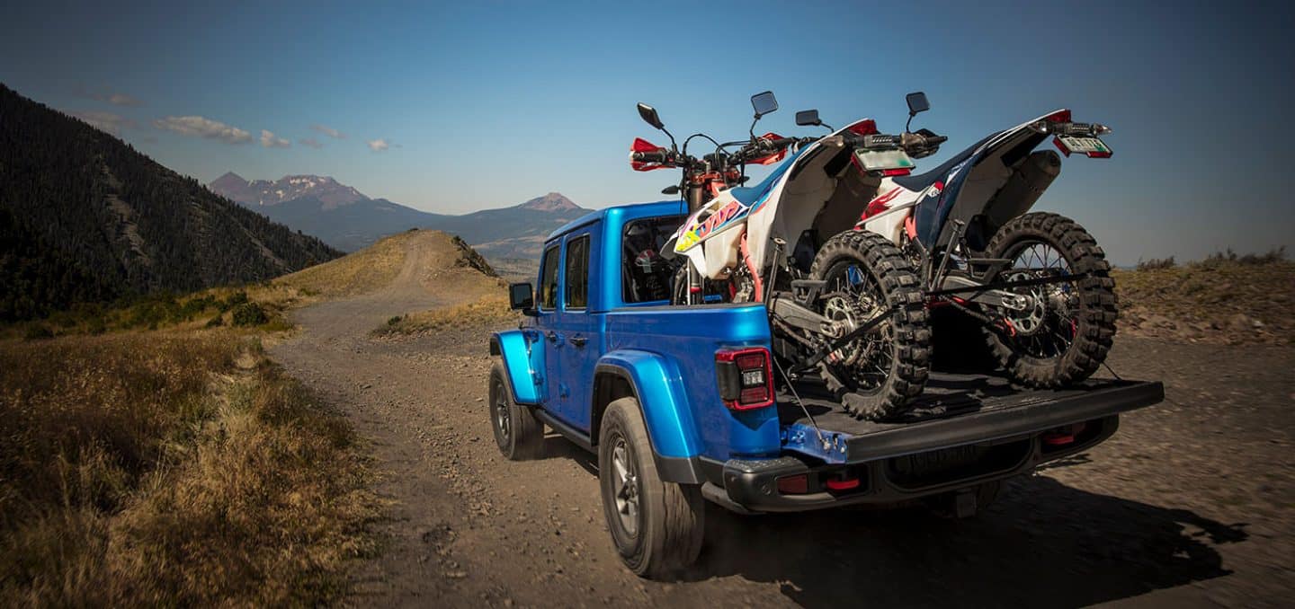 Display The rear of a blue 2025 Jeep Gladiator Rubicon X being driven on a trail off-road, with its tailgate open and two ATVs tied down in its pickup bed.