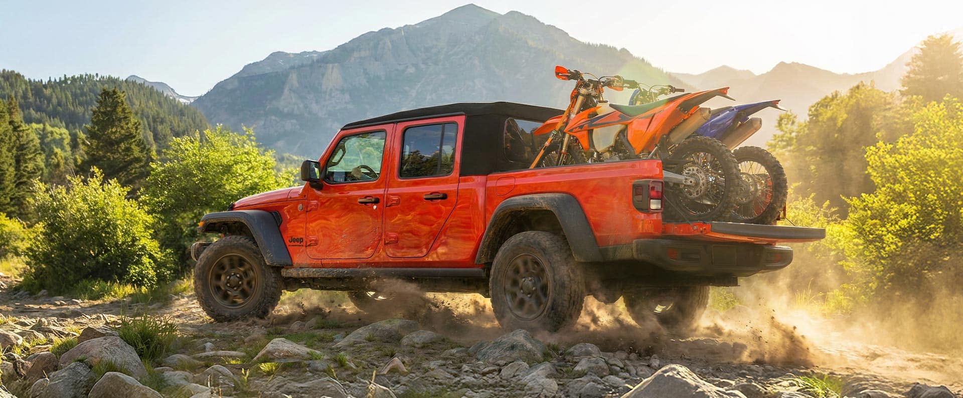A driver-side rear angle of a red 2025 Jeep Gladiator Sport S with its tailgate open, traveling on a rocky trail off-road, with two ATVs strapped inside its pickup box.