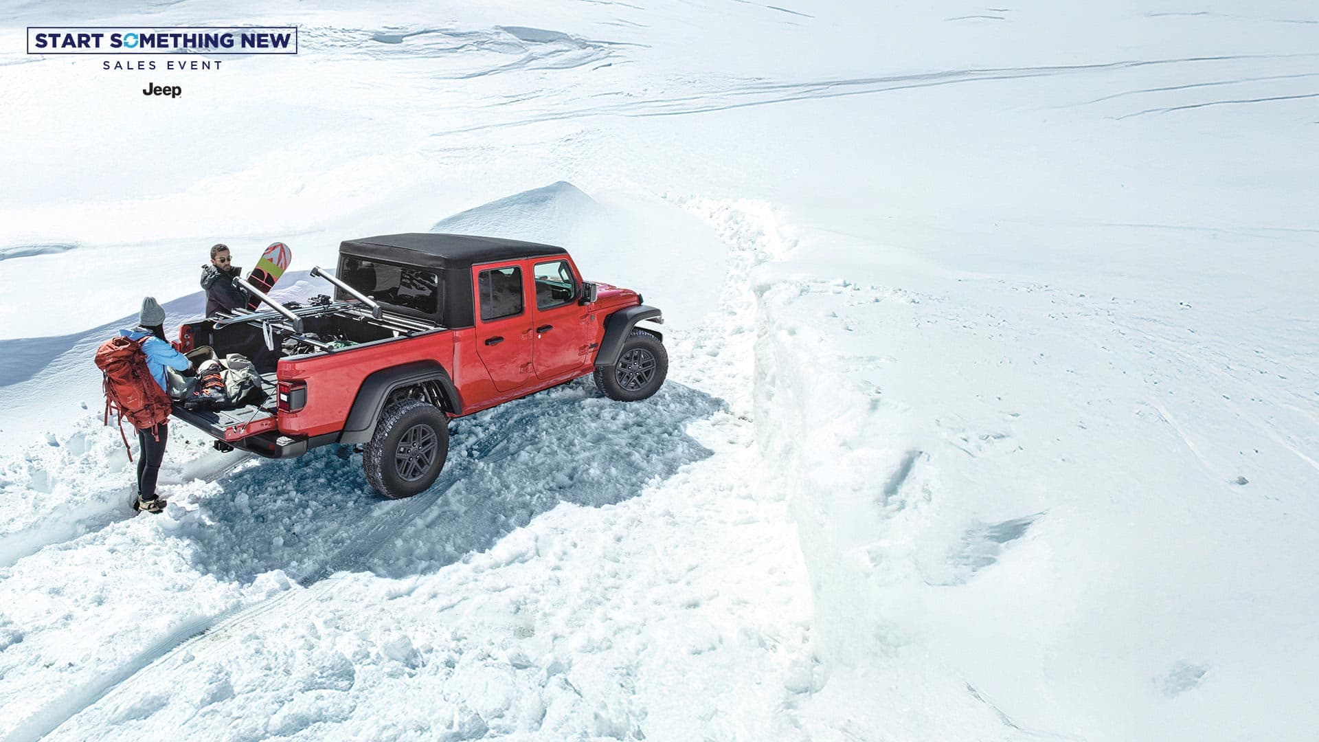 A red 2026 Jeep Gladiator Sport S with black soft top, parked in the snow with a couple removing winter sports equipment from the bed of the truck. The Start Something New Sales Event. Jeep.