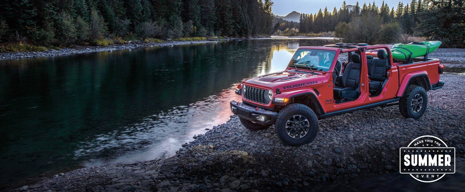 Una Jeep Gladiator Rubicon X 2025 roja con sus puertas y techo extraídos y una canoa en su plataforma, estacionada en una orilla rocosa, a la vera de un río de montaña. Evento de verano.